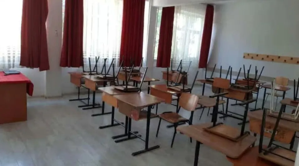 O sală de clasă cu mai multe bănci de lemn și scaune dispuse pe un podea deschisă, lângă ferestre cu perdele roșii.
