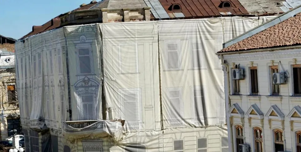 O clădire veche, parțial acoperită cu prelată albă, cu acoperiș din tablă ruginită, alături de o altă clădire cu acoperiș din țiglă și aparate de aer condiționat pe fațadă.