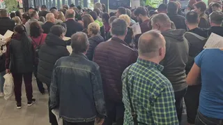 Un grup de persoane stă în picioare, cu spatele la cameră, într-un spațiu interior cu ferestre mari.