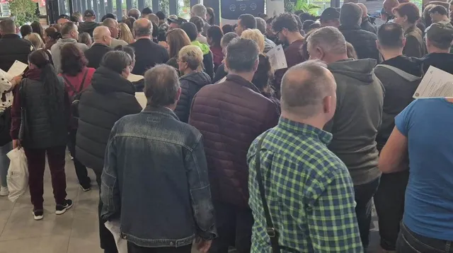 Un grup de persoane stă în picioare, cu spatele la cameră, într-un spațiu interior cu ferestre mari.