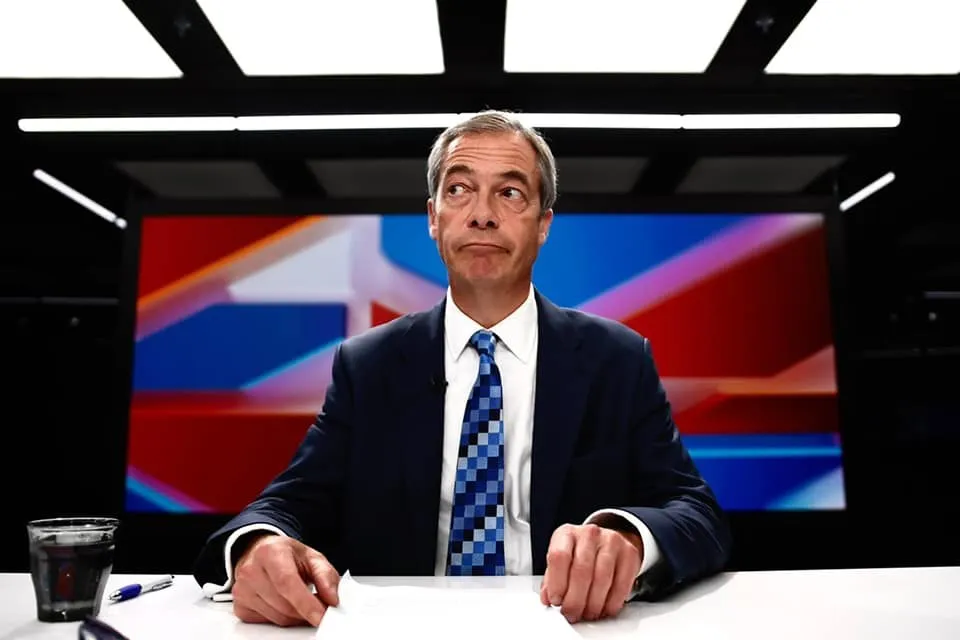 Un bărbat cu părul cărunt, purtând sacou închis la culoare și cravată albastră cu modele geometrice, stă la o masă albă în fața unui ecran colorat.