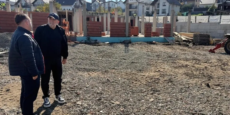 Doi bărbați stau pe un teren de construcții cu piloni de beton și stive de cărămizi roșii în fundal.
