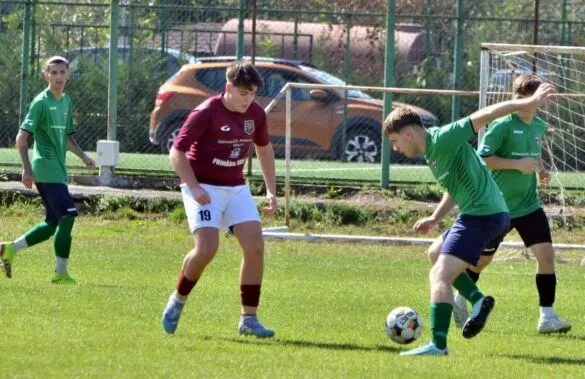 Patru jucători de fotbal sunt pe un teren de iarbă verde, fiind unul purtând o echipă de culoare roșie și alții în echipamente verzi.
