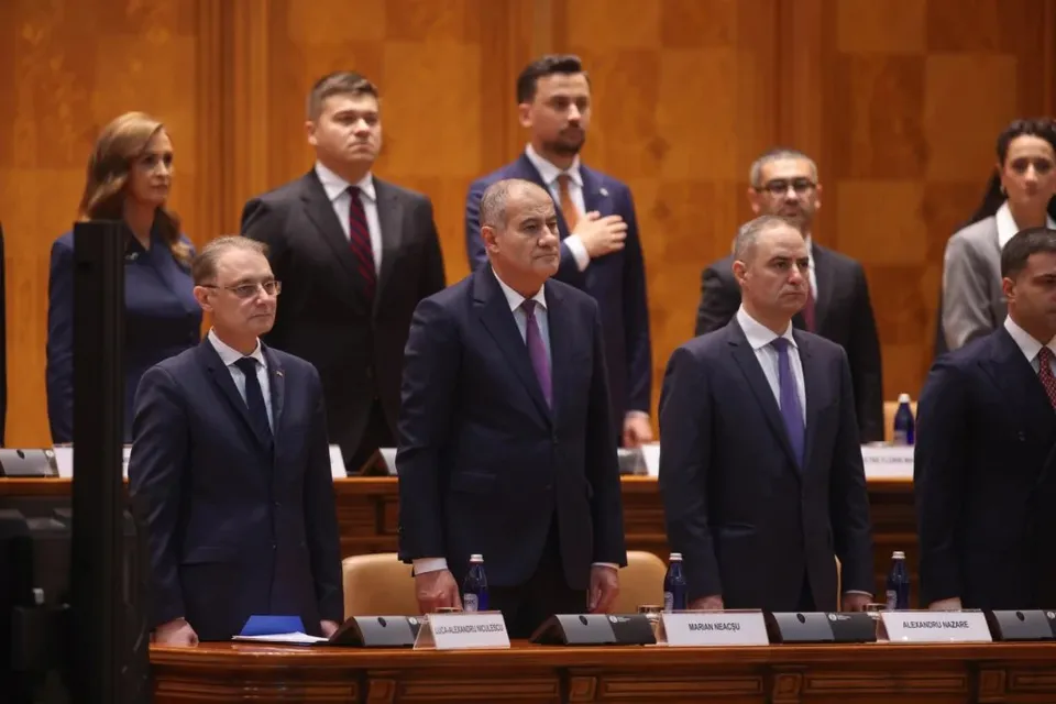 Un grup de bărbați și o femeie în costume oficiale stau în picioare în fața unor mese de lemn într-o încăpere cu panouri de lemn.