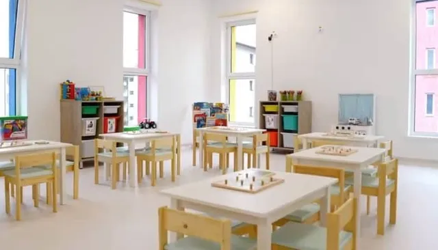 O cameră luminoasă cu mese și scaune de lemn mic, având rafturi cu jucării și materiale colorate dispuse pe margini.