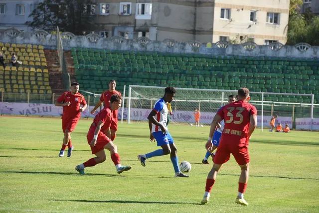 Cinci jucători de fotbal în echipamente roșii și unul în echipament albastru și alb sunt pe un teren de gazon verde.