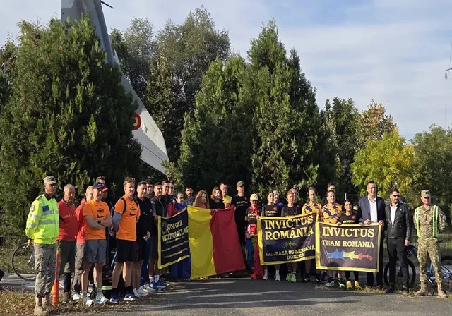 Un grup de persoane stă în rând pe un drum asfaltat, purtând haine sportive sau uniforme, lângă câteva steaguri colorate și în spatele unor copaci verzi.