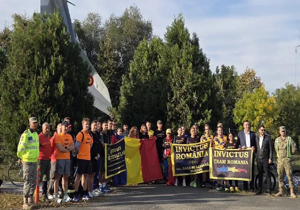 Un grup de persoane stă în rând pe un drum asfaltat, purtând haine sportive sau uniforme, lângă câteva steaguri colorate și în spatele unor copaci verzi.