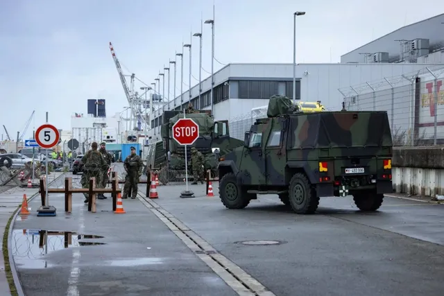 Vehicule militare de culoare verde și maro circulă pe un drum asfaltat lângă clădiri industriale și stâlpi de iluminat sub un cer înnorat.