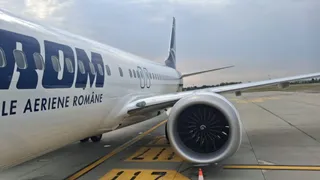 Un avion alb cu inscripția parțială TAROM și textul LE AERIENE ROMANE este parcat pe o pista de aeroport lângă un motor de turbină.