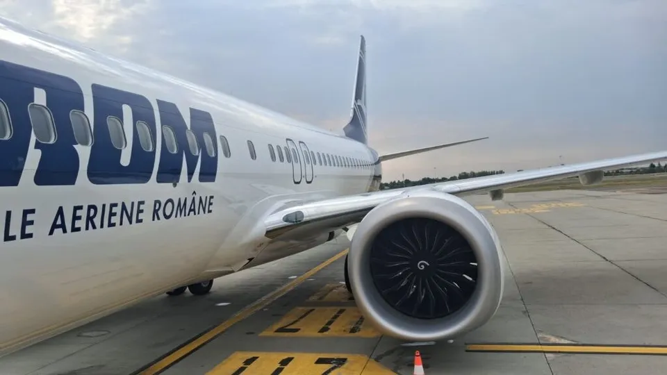 Un avion alb cu inscripția parțială TAROM și textul LE AERIENE ROMANE este parcat pe o pista de aeroport lângă un motor de turbină.