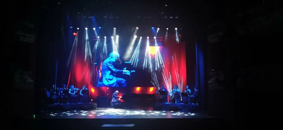 Un artist cântă la chitară pe o scenă iluminată de lumini albastre și roșii, în fața unui grup de muzicieni.
