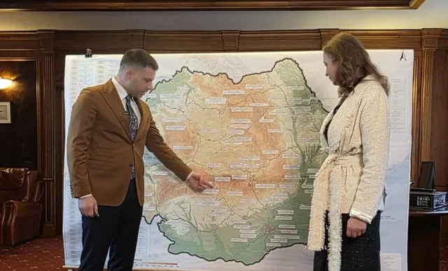 Un bărbat în costum maro și o femeie în sacou cremă stând și arătând spre o hartă a României afișată pe un panou.
