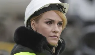 O femeie cu părul blond, purtând o cască de protecție albă și o haină cu guler de blană neagră, privește spre lateral.