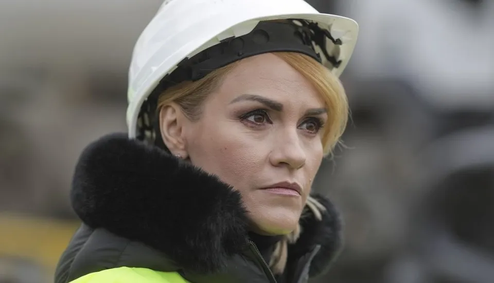O femeie cu părul blond, purtând o cască de protecție albă și o haină cu guler de blană neagră, privește spre lateral.