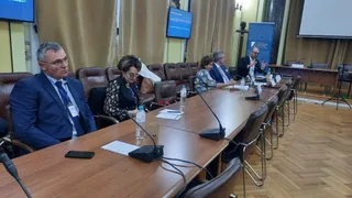 Oameni stau la o masă lungă de conferință cu microfoane, documente și sticle de apă într-o încăpere cu perdele și un ecran de proiecție.