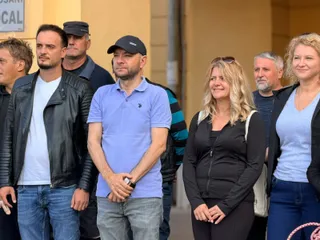 Un grup de persoane stă în picioare pe un trotuar lângă un perete galben, purtând haine casual precum tricouri, jachete și blugi.
