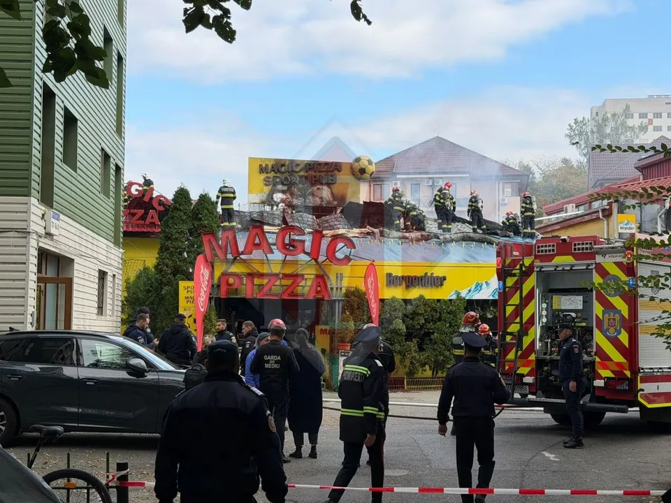 Mai multe persoane în uniforme și vehicule de intervenție sunt poziționate în fața unei clădiri cu un afiș de tip pizzerie sub un cer înnorat.