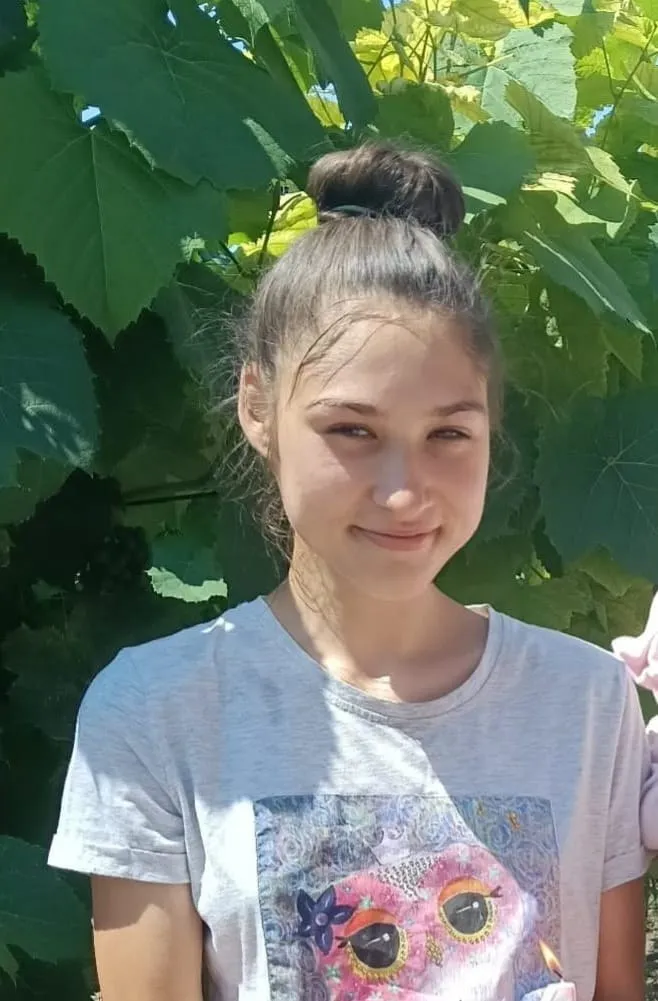 O adolescentă cu părul prins într-un coco stă în fața unor frunze verzi de viță de vie.