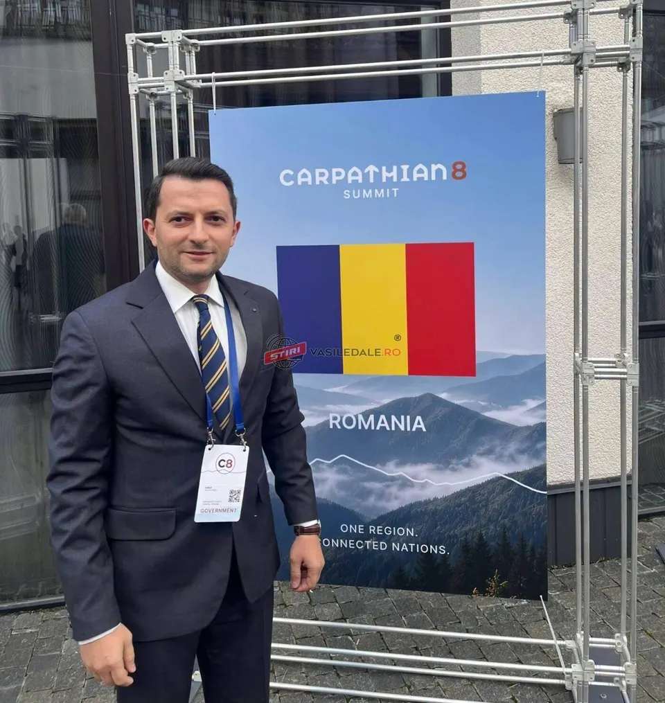 Un bărbat într-un costum închis la culoare și cravată stă în picioare lângă un panou publicitar albastru cu text și un peisaj montan.