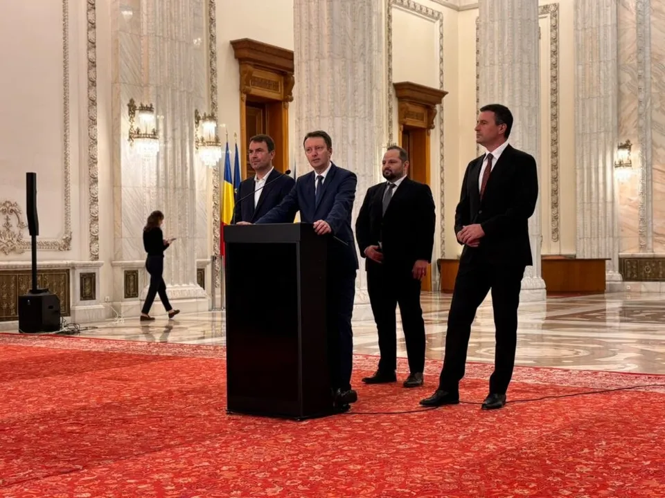 Patru bărbați în costume în plantea unui spațiu interior cu coloane înalte și un covor roșu decorat.