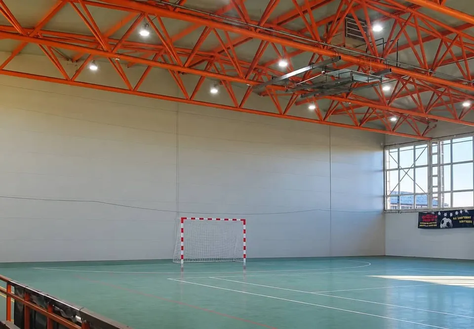 O sală de sport cu pardoseală verde, un gol de fotbal cu plasa albă și o structură de tavan metalică portocalie cu lumini suspendate.