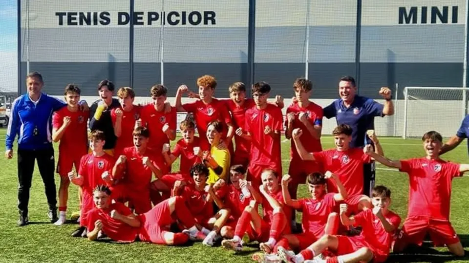 Un grup de tineri sportivi îmbrăcați în echipamente roșii posează cu brațele ridicate pe un teren de fotbal verde în fața unui gard.
