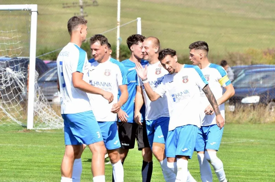Cinci jucători de fotbal în echipamente albe și albastre sărbătoresc pe un teren de gazon verde.