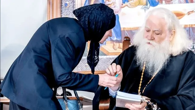 Un bărbat cu barbă albă și păr lung poartă haine negre și ține un caiet, în timp ce o femeie cu un sicliu negru se apleacă spre el.