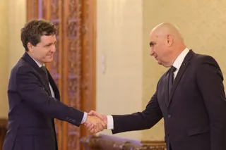 Doi bărbați în costume închise de culoare stau în picioare și se strâng pe mână într-o încăpere cu un ușă de lemn decorată în fundal.