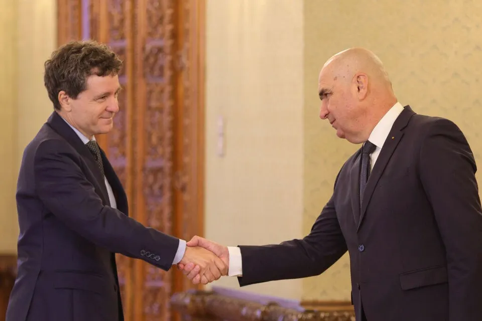 Doi bărbați în costume închise de culoare stau în picioare și se strâng pe mână într-o încăpere cu un ușă de lemn decorată în fundal.