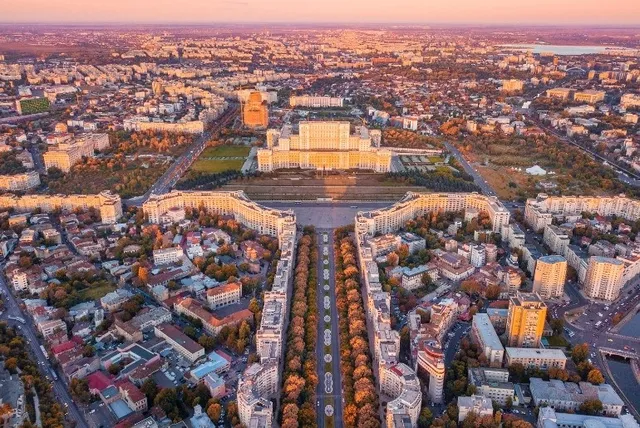 Vedere aeriană a Palatului Parlamentului din București și a bulevardului Unirii, înconjurate de clădiri și vegetație, la apus.