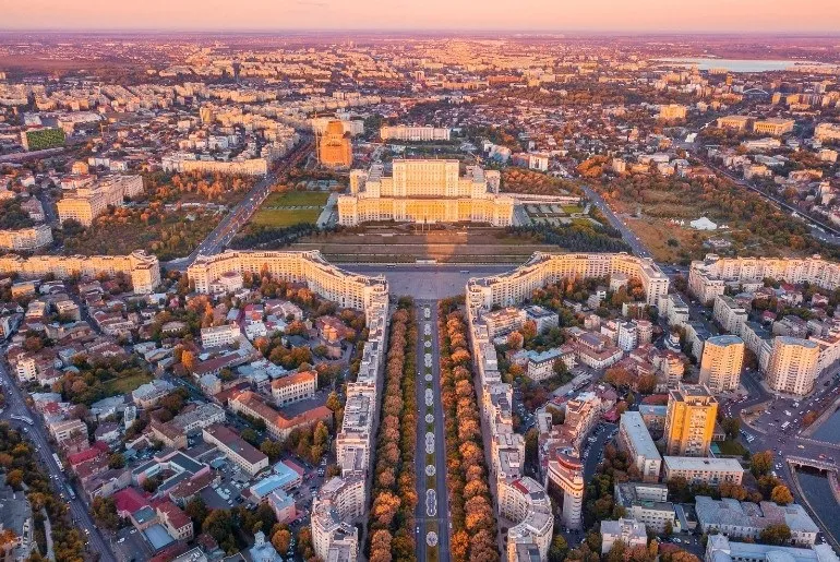 Vedere aeriană a Palatului Parlamentului din București și a bulevardului Unirii, înconjurate de clădiri și vegetație, la apus.