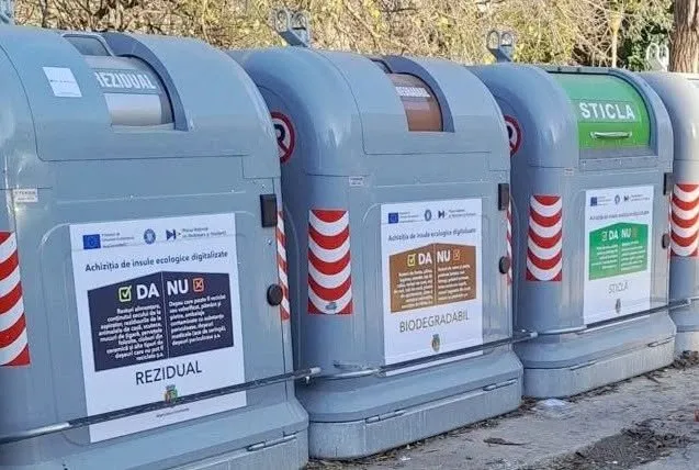 Un rând de containere gri pentru deșeuri, marcate cu etichete pentru rezidual, biodegradabil și sticlă, este situat pe un teren pietruit.