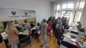 Oamenii stau la mese cu computere și documente într-o încăpere luminoasă cu ferestre mari și un panou publicitar pe perete.