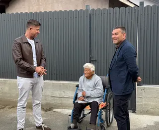 Un bătrân cu părul alb stă într-un scaun cu rotile între doi bărbați care stau în picioare lângă o gardă metalică gri.