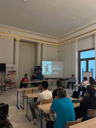 Oamenii stau la mese în fața unui ecran de proiecție care afișează un text și o imagine într-o încăpere cu coloane albe.