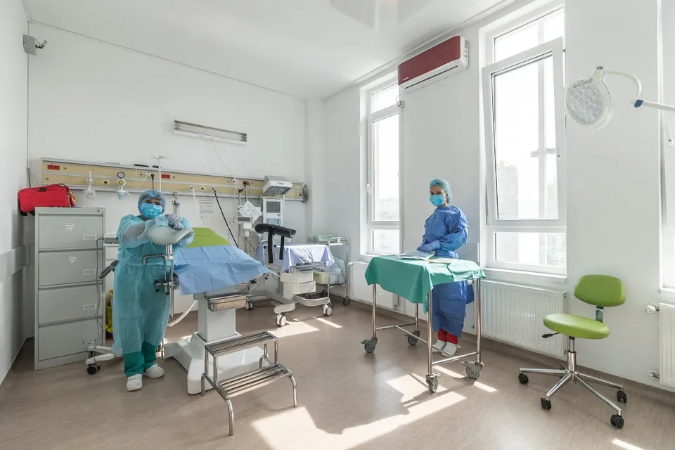 Două persoane în echipament medical albastru și măști se află într-o cameră de spital luminoasă cu echipamente medicale și ferestre mari.