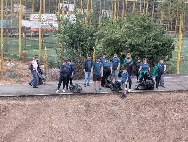 Un grup de oameni stă pe un drum asfaltat lângă saci negri de gunoi, în fața unor tufișuri verzi și a unui teren de sport împrejmuit.