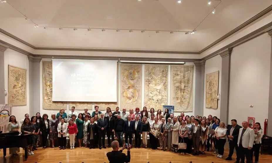 Un grup numeros de persoane stă în rânduri pe un podea de lemn în fața unui ecran alb într-o sală de expoziții iluminată.
