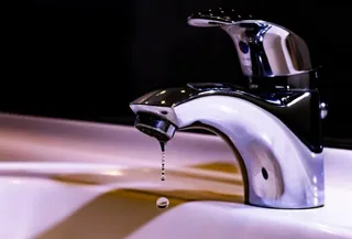 Un robinet metalic cromat lasă să cadă o picătură de apă într-un chiuvetă albă sub un fundal întunecat.