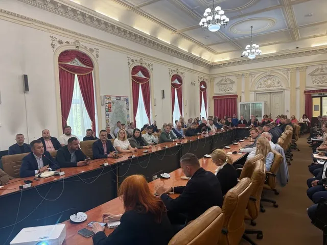 Un grup de persoane stă la mese lungi în interiorul unei săli de conferințe decorate cu ferestre înalte și lustre suspendate.