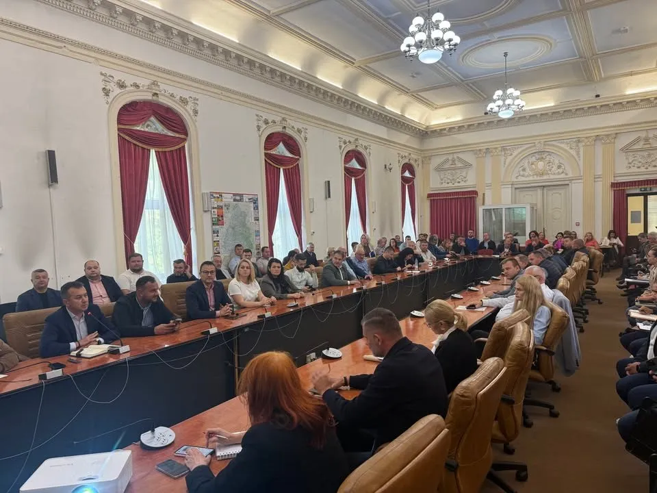 Un grup de persoane stă la mese lungi în interiorul unei săli de conferințe decorate cu ferestre înalte și lustre suspendate.