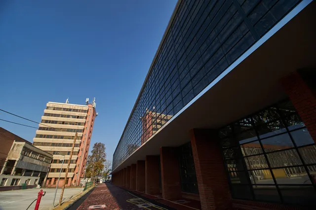 O clădire modernă cu fațadă de sticlă se întinde pe o suprafață mare, lângă un bloc de apartamente înalt sub un cer senin.