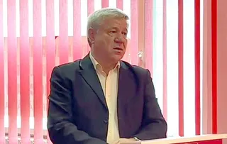 Un bărbat cu părul scurt și cămașă deschisă poartă un sacou închis la culoare stând în fața unor lămpi verticale roz și albe.