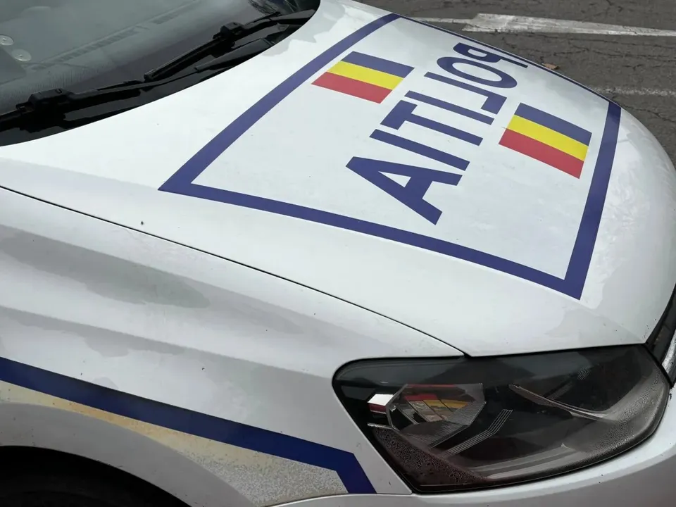 Capota unui autovehicul alb prezintă inscripția POLIȚIA și două dreptunghiuri colorate cu galben, roșu și albastru.