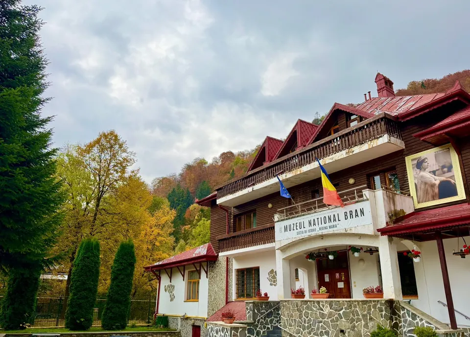 O clădire cu acoperiș roșu și un semn pe care scrie Muzeul Național Bran este înconjurată de copaci sub un cer înnorat.