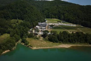 O vedere aeriană prezintă un lac cu apă verde, o zonă de câmpie și câteva clădiri situate la poalele unui munte împădurit.