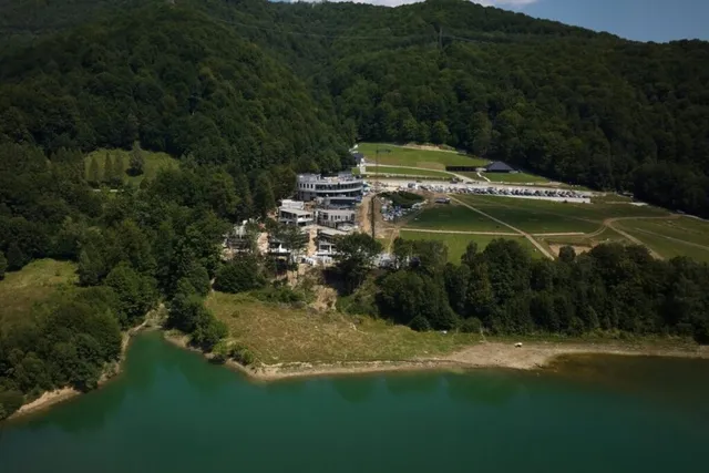 O vedere aeriană prezintă un lac cu apă verde, o zonă de câmpie și câteva clădiri situate la poalele unui munte împădurit.