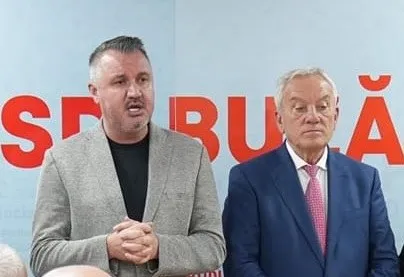 Un bărbat cu sacou gri vorbește, în timp ce un alt bărbat cu sacou albastru și cravată roz stă lângă el.